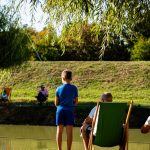 Young boy fishing by a pond on a sunny day, surrounded by trees and relaxed people.